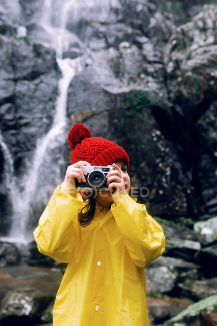 Adolescente anônimo em capa de chuva tirando foto na câmera contra montagem áspera com cascata espumosa à luz do dia — Fotografia de Stock