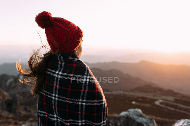 Обратный вид на неузнаваемого туриста в одеяле, стоящего на камне и наблюдающего удивительные пейзажи горной долины в солнечный день — стоковое фото