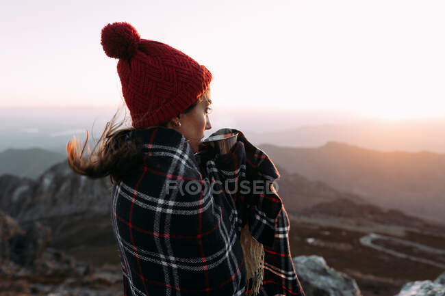 Vista lateral del excursionista en manta bebiendo café caliente y de pie sobre piedra y observando paisajes increíbles del valle de las tierras altas en un día soleado - foto de stock