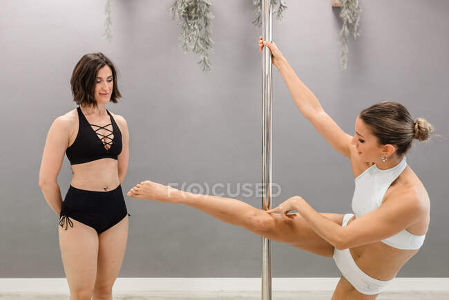 Jeune professeur en tenue de pole dance debout avec la jambe et le bras levés montrant la position du corps à l'athlète féminine en studio — Photo de stock