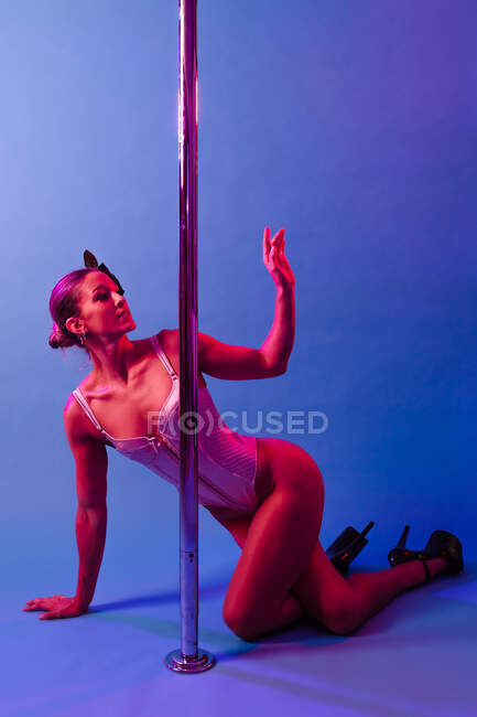 Молода витончена жінка-спортсменка в боді-костюмі і високих підборах танцює з схрещеними ногами біля металевого полюса, дивлячись на фіолетовий фон — стокове фото