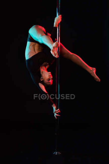 Young graceful female athlete showing inverted pose on metal pole during workout on black background — Stock Photo