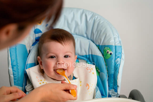 Adorable happy kid wearing bib sitting in stroller and getting fed with sweet baby food by mother — Stock Photo