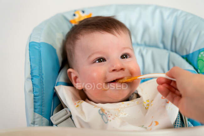 Adorable niño feliz con babero sentado en el cochecito y siendo alimentado con comida dulce para bebés por la madre - foto de stock