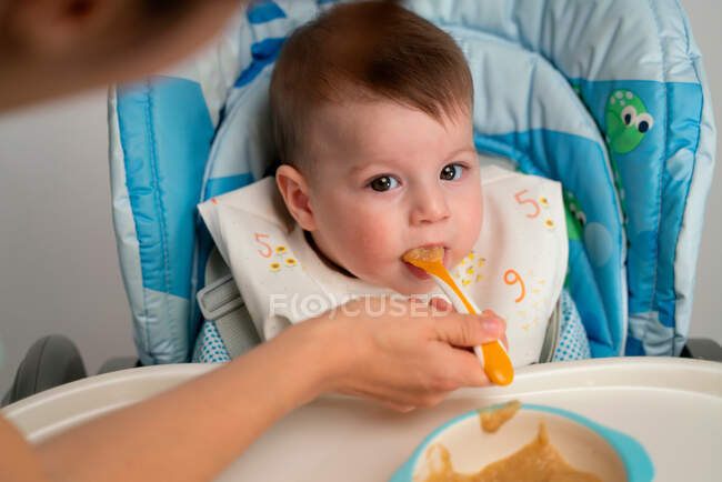 Adorable happy kid wearing bib sitting in stroller and getting fed with sweet baby food by mother — Stock Photo