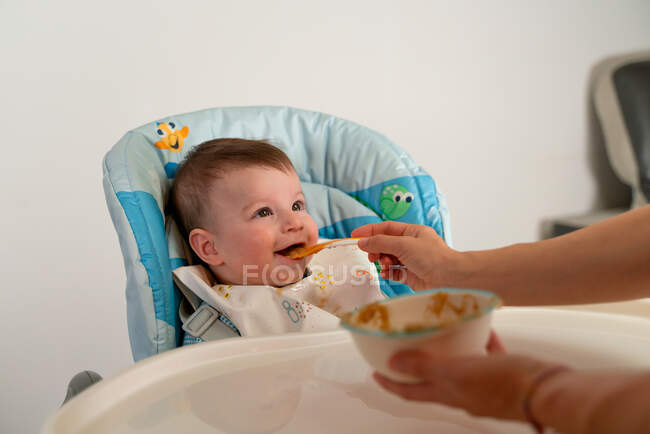 Adorable niño feliz con babero sentado en el cochecito y siendo alimentado con comida dulce para bebés por la madre - foto de stock