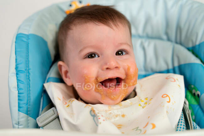 Glückliches kleines Kind im Lätzchen mit Babynahrung auf den Wangen, das auf dem Fütterungsstuhl sitzt und in die Kamera blickt — Stockfoto