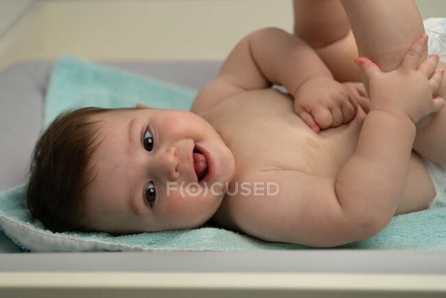 Alegre bebé en pañal acostado con las piernas levantadas en el cambiador y mirando hacia otro lado felizmente - foto de stock