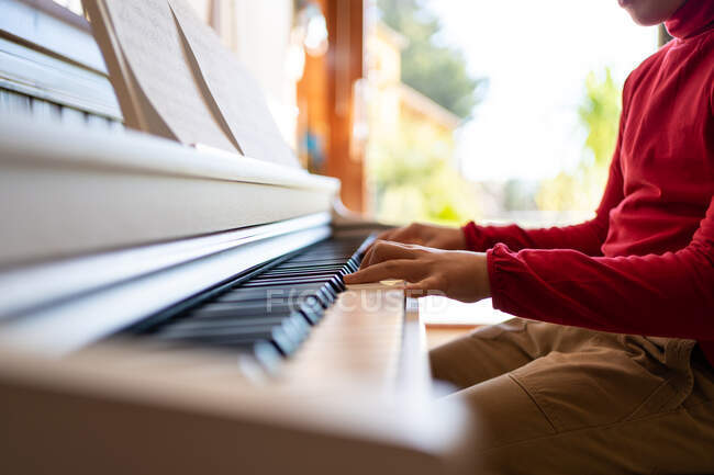 Анонимный ребенок, играющий на пианино, читая ноты и репетируя песню дома — стоковое фото