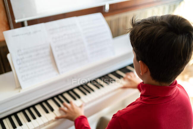 Сверху вид анонимного ребенка, играющего на пианино, читающего ноты и репетирующего песню дома — стоковое фото