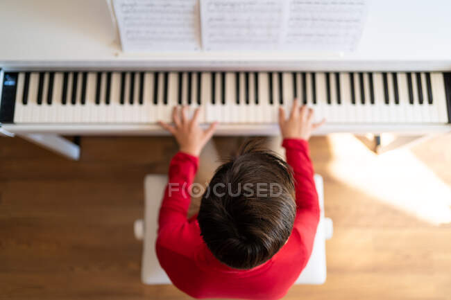 Vue de dessus du dos d'un enfant anonyme jouant du piano tout en lisant des notes et en répétant une chanson à la maison — Photo de stock