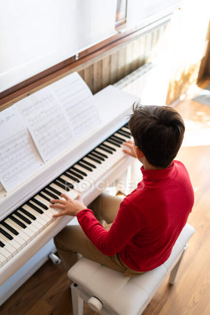 Desde arriba vista posterior de niño anónimo tocando el piano mientras lee notas y ensayando la canción en casa - foto de stock
