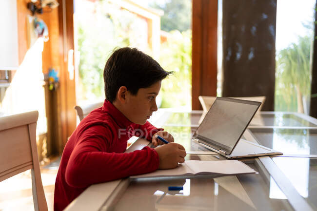 Side view of smart schoolboy sitting at table with laptop and writing in notebook while doing homework alone — Stock Photo