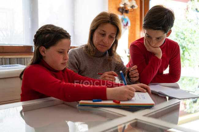 Adolescente colegiala sentada en la mesa con la madre y el hermano y escribiendo en copybook mientras hacen la tarea juntos en casa - foto de stock