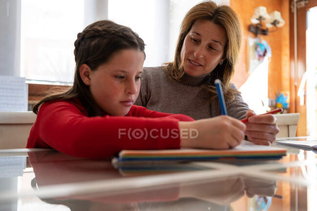 Мама помогает дочери с домашним заданием, сидя за столом с ноутбуком и учится дома — стоковое фото