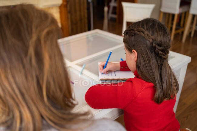 Високий кут назад невизначеної матері, яка допомагає доньці з домашнім завданням, сидячи за столом з блокнотом і навчаючись вдома — стокове фото