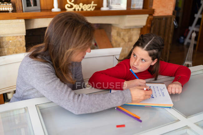 Angle élevé de la mère aider sa fille à faire ses devoirs tout en étant assis à table avec un cahier et en étudiant à la maison — Photo de stock