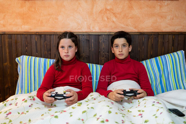 Alegre hermano adolescente y hermana sentados en la cama y jugando videojuegos mientras usa gamepads y disfruta el fin de semana en casa - foto de stock