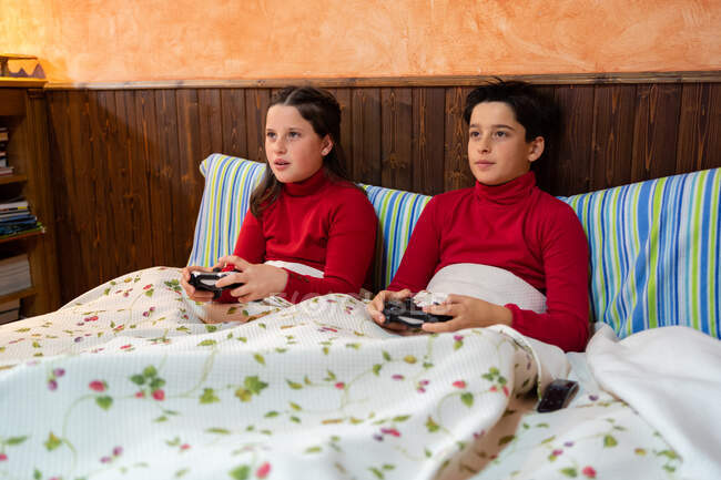 Веселые брат и сестра-подросток сидят на кровати и играют в видеоигры, используя геймпады и наслаждаясь выходными дома — стоковое фото