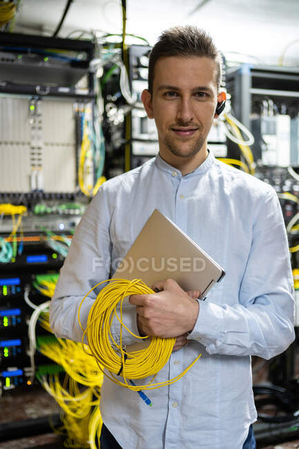 Positive man in wireless headset standing with netbook and cables in server room — Stock Photo