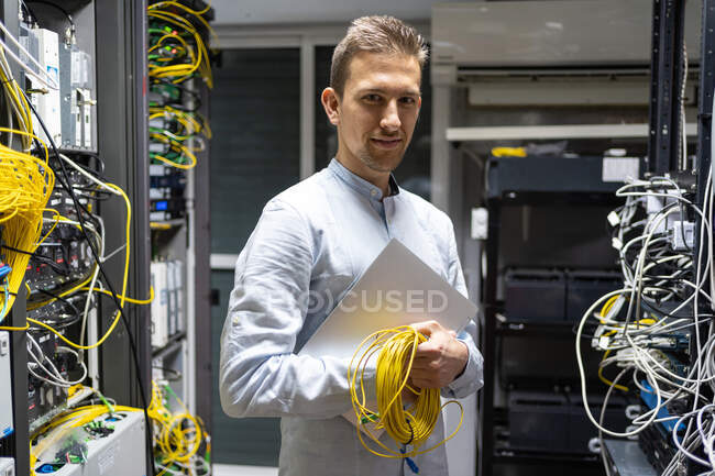 Positive man in wireless headset standing with netbook and cables in server room — Stock Photo