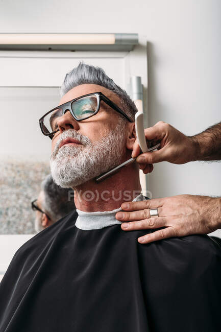 Low angle of crop unrecognizable male barber shaving gray beard of middle aged stylish client in eyeglasses sitting near mirror in hairdressing salon — Stock Photo