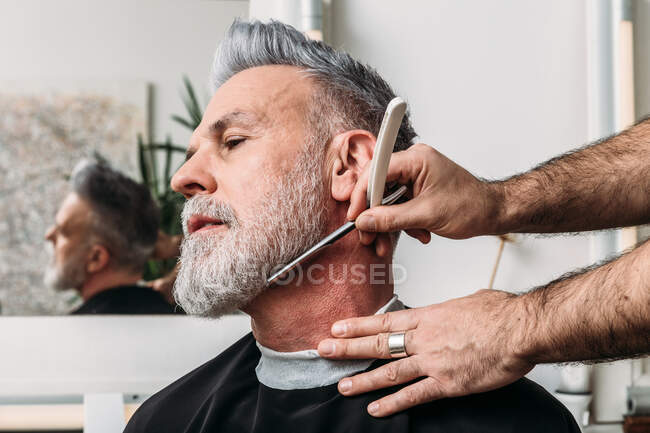 Низкий угол урожая неузнаваемый мужчина парикмахер бреет седую бороду средневекового стильного клиента в очках, сидящих возле зеркала в парикмахерской — стоковое фото