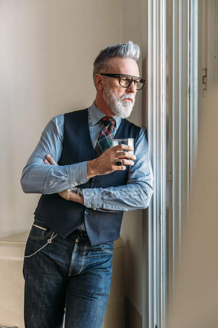 Homme élégant d'âge moyen confiant aux cheveux gris et à la barbe en tenue tendance et aux lunettes penchées sur le mur et regardant par la fenêtre tout en buvant du whisky dans l'appartement — Photo de stock