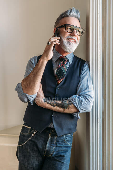 Conteúdo maduro masculino executivo em elegante desgaste e óculos falando no celular enquanto se inclina na parede e olha para longe na construção — Fotografia de Stock