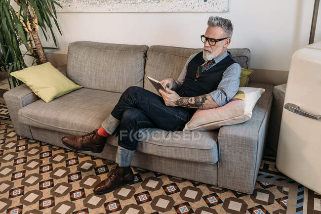 Trendy uomo d'affari di mezza età navigazione internet su tablet mentre seduto con le gambe incrociate sul divano in casa — Foto stock