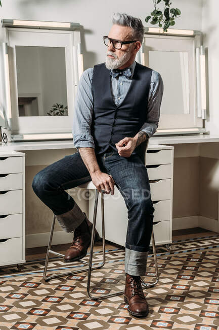 Trendy maturo uomo d'affari hipster in occhiali e vestiti alla moda guardando lontano sulla sedia in casa — Foto stock