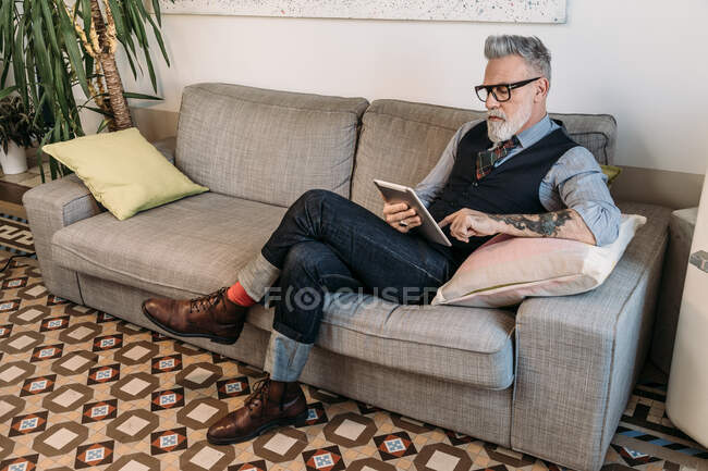Moderno hombre de negocios de mediana edad navegando por Internet en la tableta mientras está sentado con las piernas cruzadas en el sofá en casa - foto de stock
