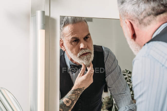 Crop serious middle aged male executive with tattoo touching gray beard while looking in mirror in house — Stock Photo