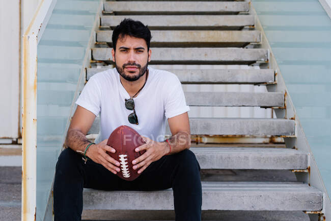 Confident young bearded ethnic male in white t shirt sitting on stairs on street and looking at camera with rugby ball in hands — Stock Photo