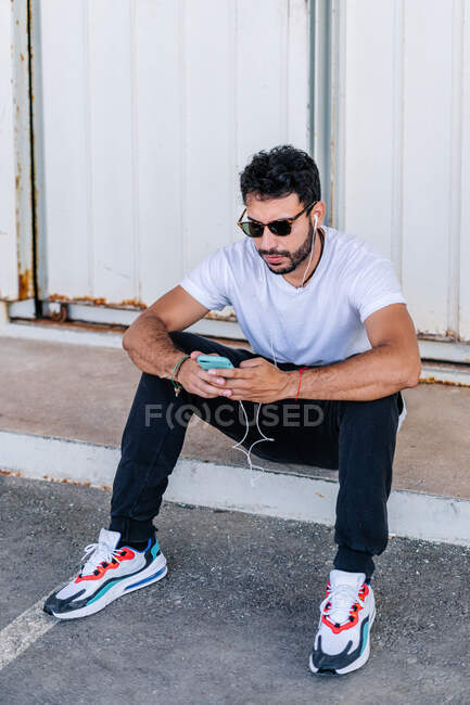 Full body serious bearded male in casual wear and sunglasses browsing modern mobile phone and listening to music via earphones while sitting on asphalt sidewalk on street — Stock Photo