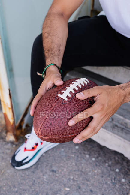 Irreconhecível jovem étnico masculino segurando bola de rugby enquanto ele está sentado no dia ensolarado — Fotografia de Stock