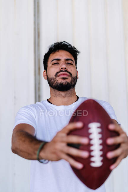 Auto assicurato giovane maschio etnico barbuto in t shirt bianca con pelle di maiale mentre giocava a football americano nella giornata di sole — Foto stock