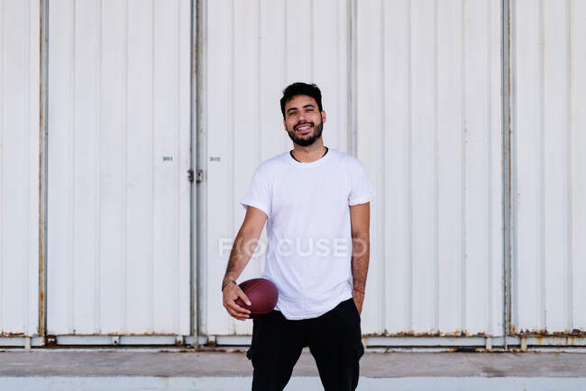 Самовпевнений молодий бородатий етнічний чоловік у білій футболці з піхвою, граючи в американський футбол у сонячний день. — стокове фото