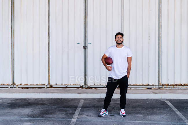 Self assured young bearded ethnic male in white t shirt holding pigskin while playing American football on sunny day — Stock Photo