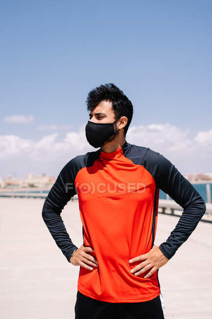 Мужчина в маске, стоящий с руками на талии в солнечном парке перед открытыми тренировками — стоковое фото