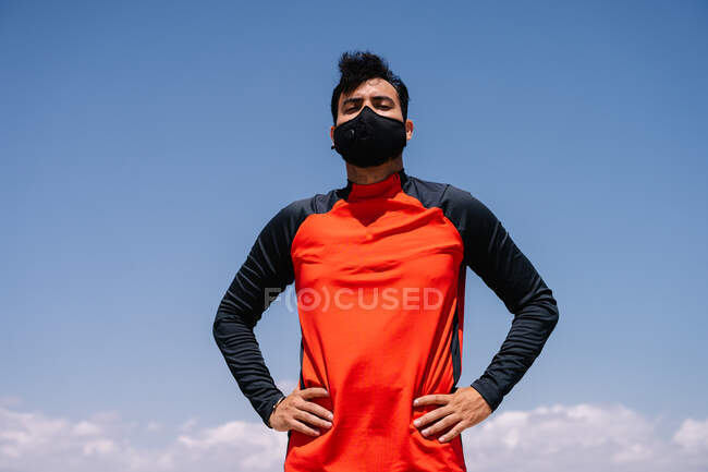 Atleta masculino em máscara de pé com as mãos na cintura no parque ensolarado antes do treinamento ao ar livre — Fotografia de Stock
