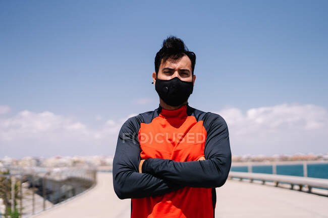 Мужчина в маске, стоящий с руками, скрещенными в солнечном парке перед открытыми тренировками — стоковое фото