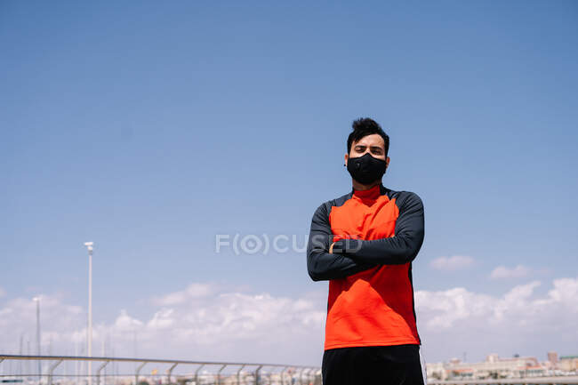Hombre atleta en máscara de pie con los brazos cruzados en el parque soleado antes del entrenamiento al aire libre - foto de stock