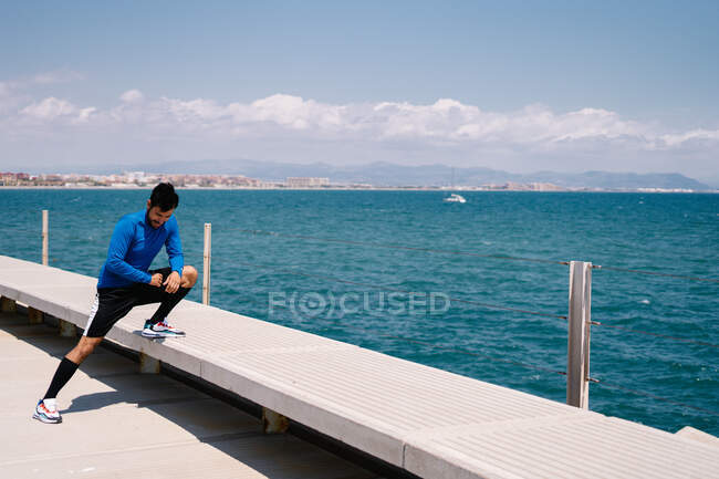 Cuerpo completo de atleta masculino atlético determinado que calienta el cuerpo mientras se prepara para el entrenamiento en el paseo marítimo - foto de stock