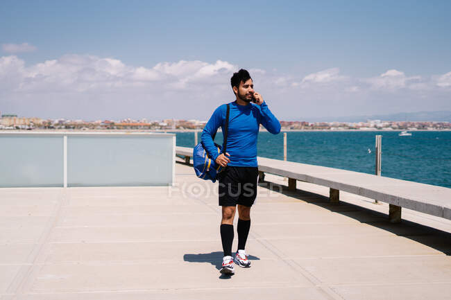 Atleta masculino sonriente teniendo conversación en el teléfono móvil después del entrenamiento en el terraplén de la ciudad en el día soleado - foto de stock