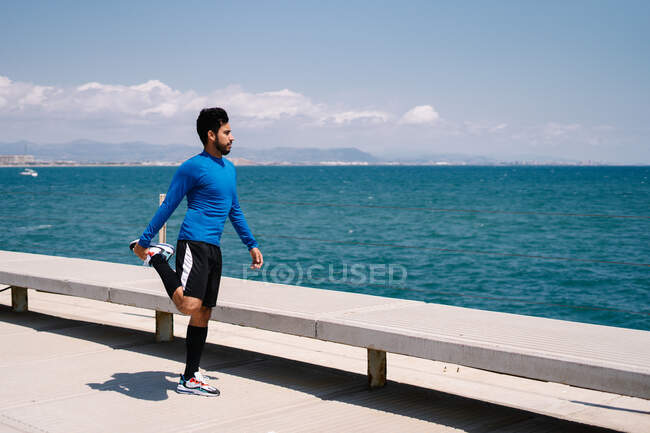 Corpo inteiro do atleta masculino atlético determinado aquecendo o corpo enquanto se prepara para o treino à beira-mar — Fotografia de Stock