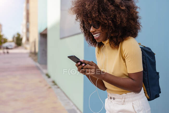 Чорна жінка з афро-волоссям слухає музику на мобільному перед синьою стіною — стокове фото