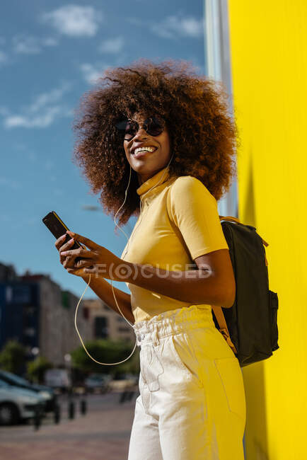 Черная женщина с афроволосами слушает музыку на мобильном телефоне перед жёлтой стеной — стоковое фото