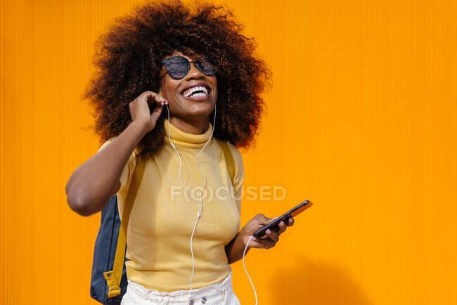 Черная женщина с афроволосами слушает музыку на мобильном телефоне перед оранжевой стеной — стоковое фото