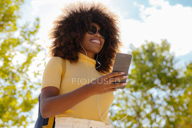 Черная женщина с афроволосами слушает музыку на мобильном телефоне с рюкзаком на спине — стоковое фото